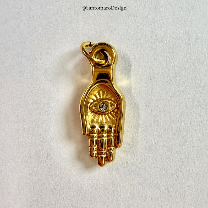 May include: A gold-coloured pendant shaped like a hand, with an eye design at the centre, set with a small, clear stone. The pendant has a loop at the top for a chain. The design is detailed and ornate.