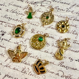 Pu&ograve; includere: Una collezione di ciondoli dorati con vari disegni, tra cui angeli, raggi di sole e una farfalla. Diversi ciondoli presentano pietre preziose verdi o nere. I ciondoli sono esposti su uno sfondo di carta vintage.