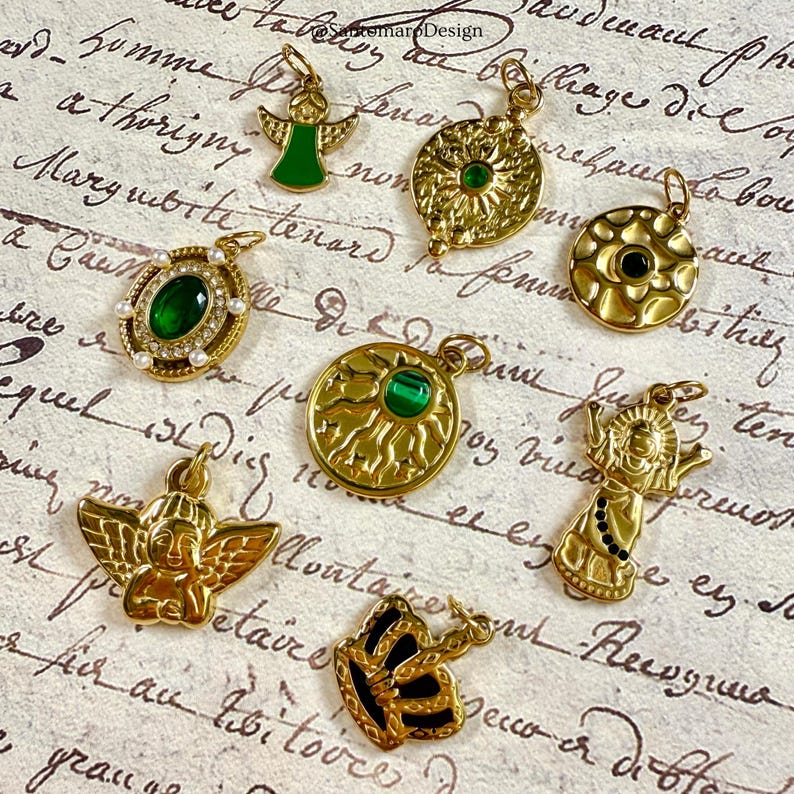 Pu&ograve; includere: Una collezione di ciondoli dorati con vari disegni, tra cui angeli, raggi di sole e una farfalla. Diversi ciondoli presentano pietre preziose verdi o nere. I ciondoli sono esposti su uno sfondo di carta vintage.