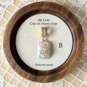 May include: A natural pearl charm with an 18K gold clip-on clasp. The pearl is rectangular with a pink rose and gold butterfly embellishment. The charm is displayed in a wooden bowl on a cream-coloured surface. The text "Natural pearl" is visible.