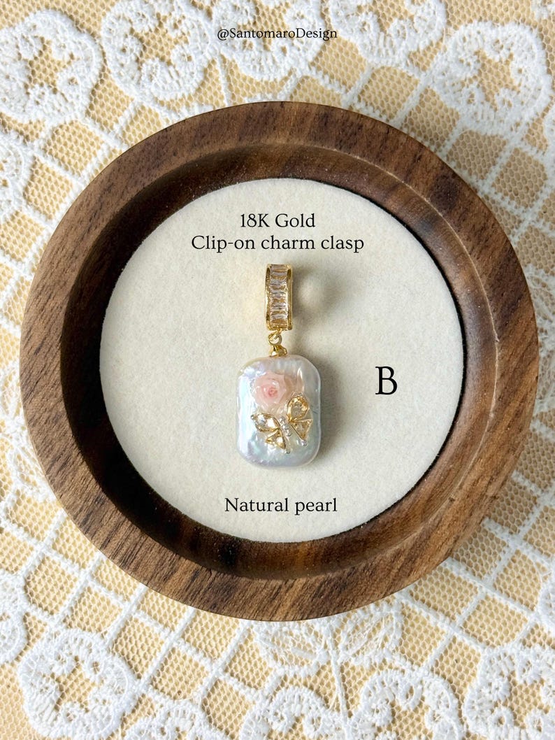 May include: A natural pearl charm with an 18K gold clip-on clasp. The pearl is rectangular with a pink rose and gold butterfly embellishment. The charm is displayed in a wooden bowl on a cream-coloured surface. The text "Natural pearl" is visible.