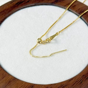 May include: Close-up of a gold-colored necklace with an adjustable chain and clasp. The necklace is on a white surface, and the background is a wooden frame. The necklace has a delicate, box chain design.