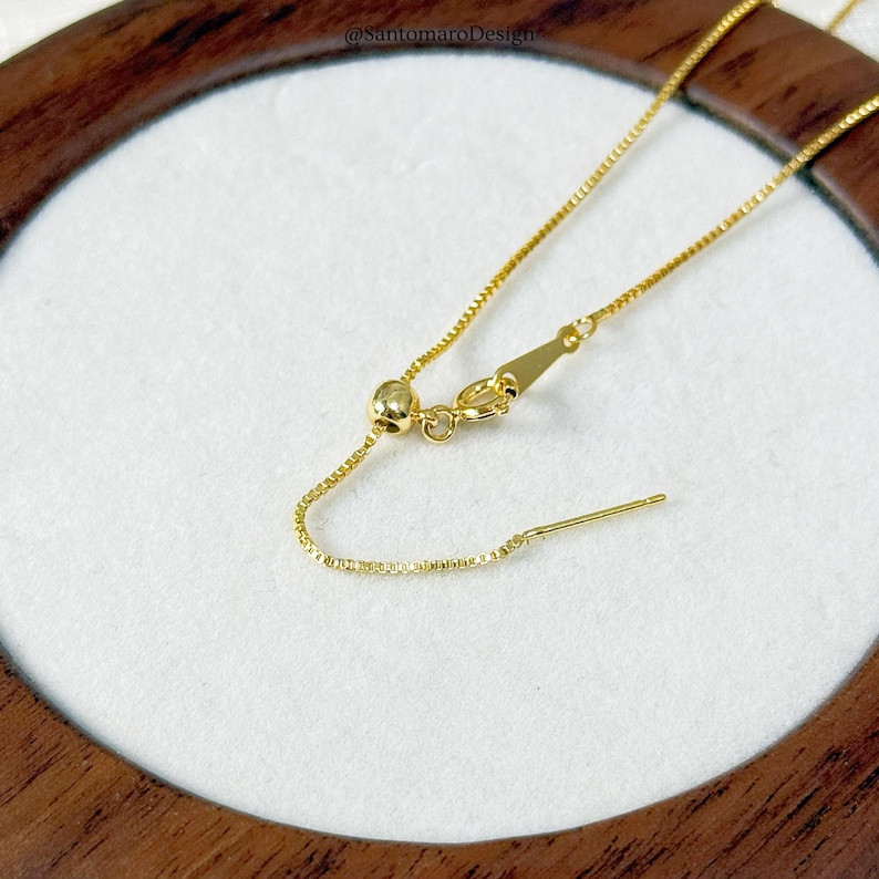 May include: Close-up of a gold-colored necklace with an adjustable chain and clasp. The necklace is on a white surface, and the background is a wooden frame. The necklace has a delicate, box chain design.