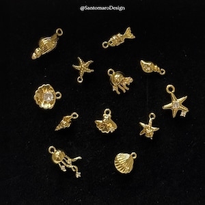 Può includere: Una collezione di ciondoli color oro in varie forme a tema marino, tra cui stelle marine, conchiglie e un pesce. Alcuni ciondoli hanno piccoli abbellimenti trasparenti. Lo sfondo è nero e i ciondoli sono disposti in modo sparso.