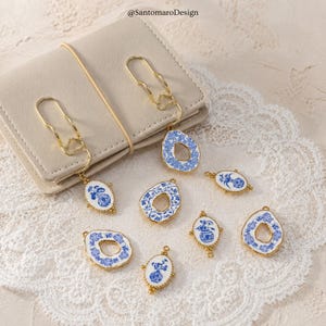 Könnte beinhalten: Eine Sammlung von blau-weißen Porzellananhängern mit goldfarbenen Metallakzenten. Die Anhänger weisen florale und abstrakte Designs auf. Einige sind an goldfarbenen Büroklammern befestigt, vor einem hellen Notizbuch und Spitze.