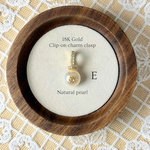 May include: A natural pearl charm with an 18K gold clip-on clasp. The charm is displayed in a wooden tray with a white interior. The text "18K Gold Clip-on charm clasp" and "Natural pearl" are visible.