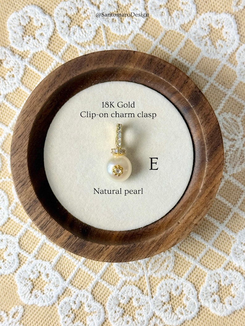 May include: A natural pearl charm with an 18K gold clip-on clasp. The charm is displayed in a wooden tray with a white interior. The text "18K Gold Clip-on charm clasp" and "Natural pearl" are visible.