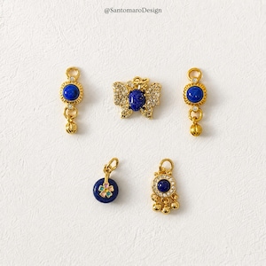 Può includere: Una collezione di ciondoli per gioielli color oro con accenti di pietre preziose blu. I ciondoli includono disegni a farfalla, rotondi e a goccia. I gioielli sono presentati su uno sfondo bianco. Il testo "@SantomaroDesign" è visibile.