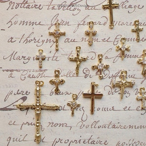 May include: A collection of gold-colored cross charms, some with clear gemstone accents. The crosses vary in size and design, with intricate details. The background features handwritten script. The text "@SantamaroDesign" is visible.