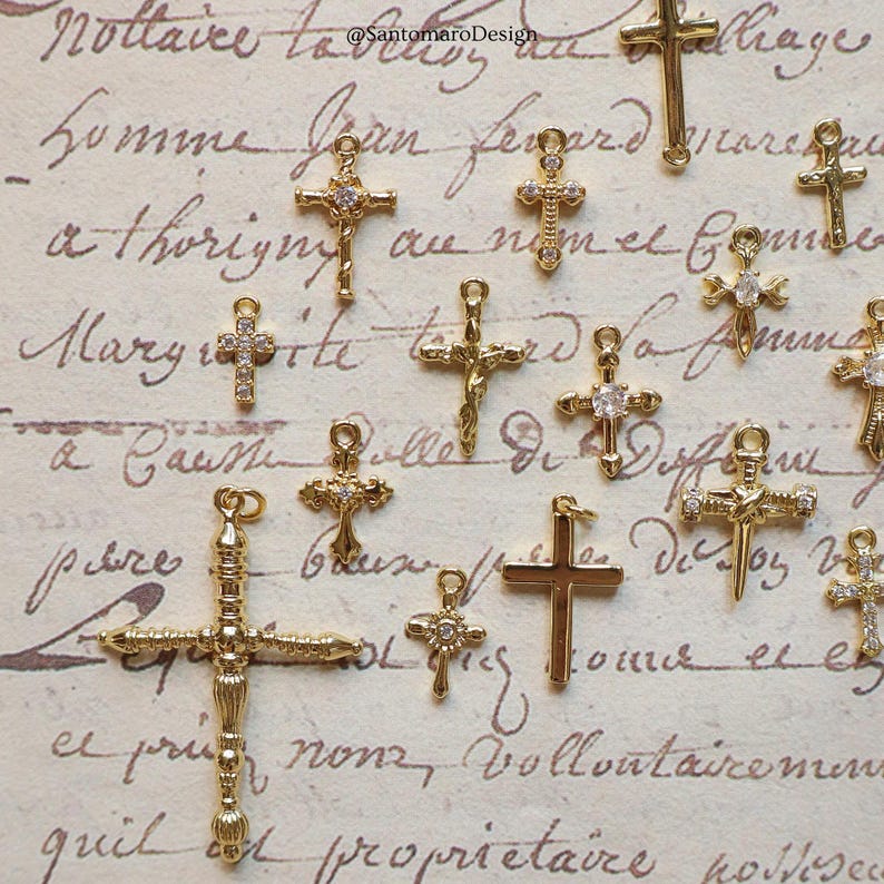May include: A collection of gold-colored cross charms, some with clear gemstone accents. The crosses vary in size and design, with intricate details. The background features handwritten script. The text "@SantamaroDesign" is visible.