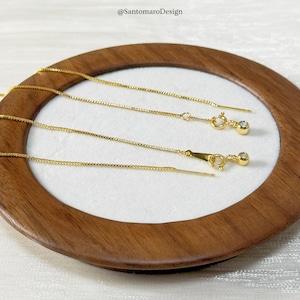 May include: Close-up of two gold-colored threader earrings with small, round, clear gemstones. The earrings are displayed on a wooden tray with a white interior. The text "@SantomaroDesign" is visible in the upper left corner.