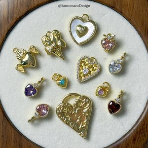 May include: A selection of gold-coloured heart-shaped charms with diverse designs, including those with gemstones, wings, and intricate patterns. Some charms feature clear, pink, and purple stones. The charms are displayed on a white surface.