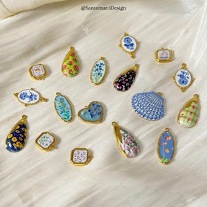 Può includere: Una collezione di ciondoli dorati con vari disegni, tra cui motivi floreali, una conchiglia e una forma di cuore. I ciondoli presentano dettagli in smalto colorato nei toni del blu, rosa, verde e giallo. L'immagine include il testo "@SantomaroDesign".