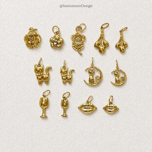 May include: A collection of gold-colored charms in various designs, including flowers, grapes, cats, wine glasses, and lips. The charms are displayed on a light-colored surface. The text "@SantamaroDesign" is visible at the top.