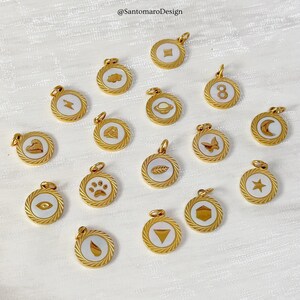 Könnte beinhalten: Eine Sammlung goldfarbener Anhänger mit verschiedenen weißen Emaille-Designs, darunter ein Blitz, ein Diamant und ein Pfotenabdruck. Jeder Anhänger hat einen seilartigen Rand und eine kleine Schlaufe für eine Kette.
