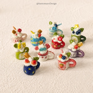 Peut inclure: Une collection de tasses miniatures en céramique, chacune décorée de crème fouettée colorée, de fruits et de motifs floraux. Les tasses présentent diverses couleurs, dont le rose, le bleu, le vert et le rouge, avec des détails d'étoiles et de cœurs. Le texte "@SantomaroDesign" est visible.