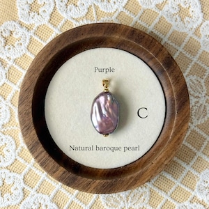 May include: A natural baroque pearl pendant displayed in a wooden holder. The purple pearl hangs from a gold-coloured bail. The text "Purple", "C", and "Natural baroque pearl" are visible.