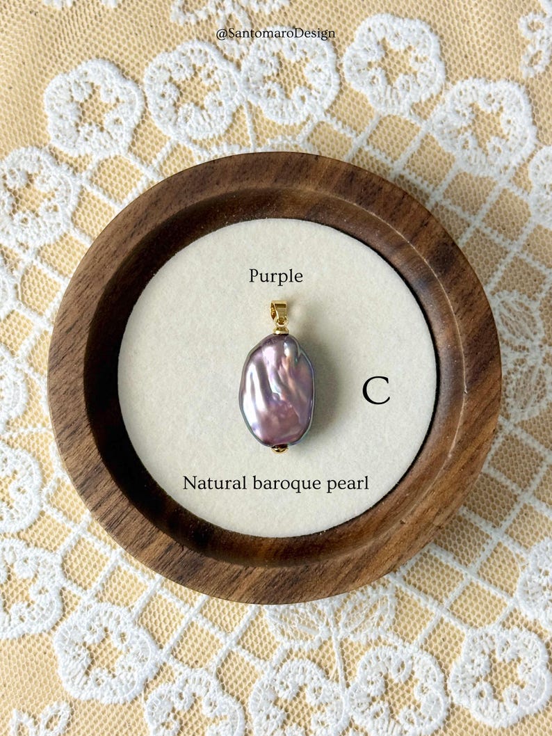 May include: A natural baroque pearl pendant displayed in a wooden holder. The purple pearl hangs from a gold-coloured bail. The text "Purple", "C", and "Natural baroque pearl" are visible.