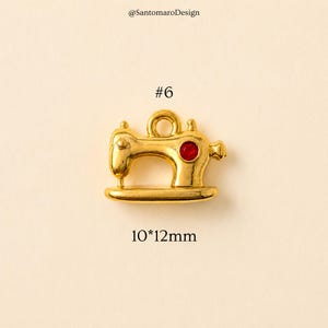 May include: A gold-colored sewing machine charm with a red gem, measuring approximately 0.4 x 0.5 inches. The charm has a loop at the top and is labeled with the number "#6". The text "@SantomaroDesign" is at the top.