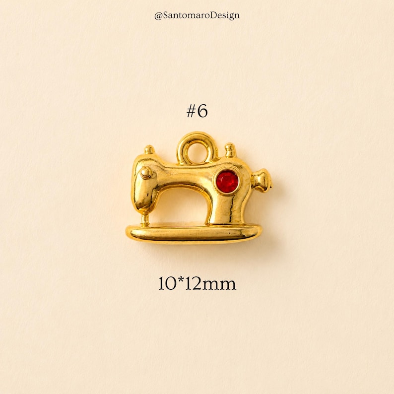 May include: A gold-colored sewing machine charm with a red gem, measuring approximately 0.4 x 0.5 inches. The charm has a loop at the top and is labeled with the number "#6". The text "@SantomaroDesign" is at the top.