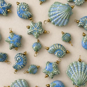 Op de afbeelding: Een verzameling keramische bedels in blauwe en groene tinten, gevormd als schelpen en zeesterren. Elk stuk heeft een glanzende afwerking en is voorzien van kleine goudkleurige metalen kralen en ringetjes. De tekst @SantomaroDesign is bovenaan zichtbaar.