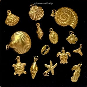 Puede incluir: Una colección de colgantes dorados con forma de conchas marinas, incluyendo una estrella de mar, un caballito de mar y una tortuga. Los colgantes son detallados y tienen un acabado pulido. Los colgantes están dispuestos sobre una superficie negra.