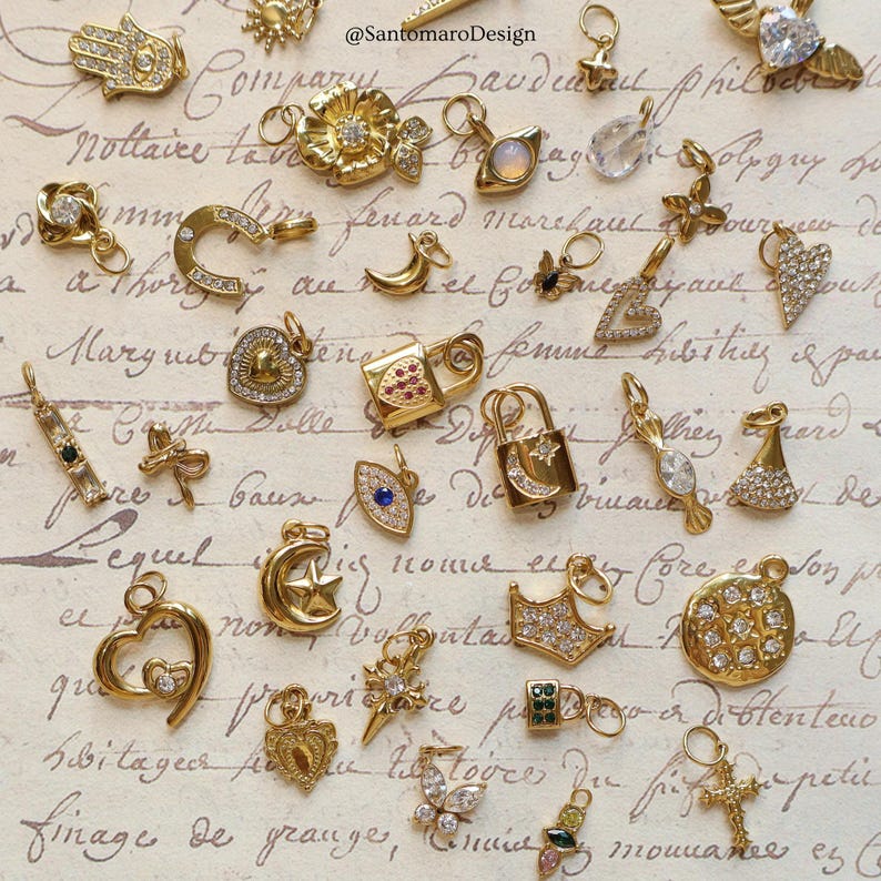 May include: A collection of gold-toned charms with various designs, including hearts, crosses, moons, stars, and floral motifs. Some charms are embellished with clear and colored gemstones. The charms are displayed on a vintage paper background.