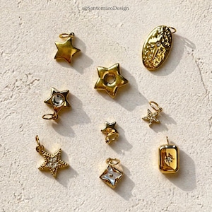 Peut inclure: Une collection de breloques et de pendentifs dorés en forme d'étoile. Certains charms présentent des cristaux transparents ou une incrustation de nacre. Les charms varient en taille et en design, avec un pendentif rectangulaire en bas à droite. Le texte "@SantamaroDesign" est inclus.