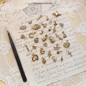 May include: A collection of gold-toned charms with various designs, including hearts, crosses, moons, and locks, arranged on a vintage paper background. A pair of black tweezers is on the left. The charms are embellished with crystals and other decorative elements.