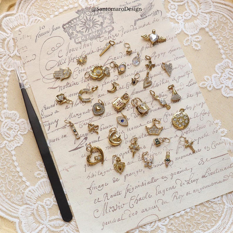 May include: A collection of gold-toned charms with various designs, including hearts, crosses, moons, and locks, arranged on a vintage paper background. A pair of black tweezers is on the left. The charms are embellished with crystals and other decorative elements.