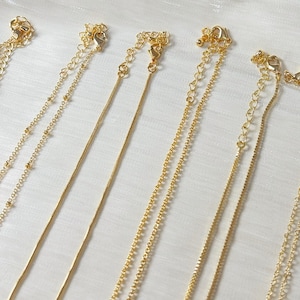 Op de afbeelding: Close-up van verschillende goudkleurige kettingen met diverse kettingstijlen. De kettingen hebben verschillende ontwerpen, waaronder kralen-, slang- en schakelkettingen. Elke ketting heeft een sluiting en een verstelbare ketting. De achtergrond is een witte, getextureerde stof.