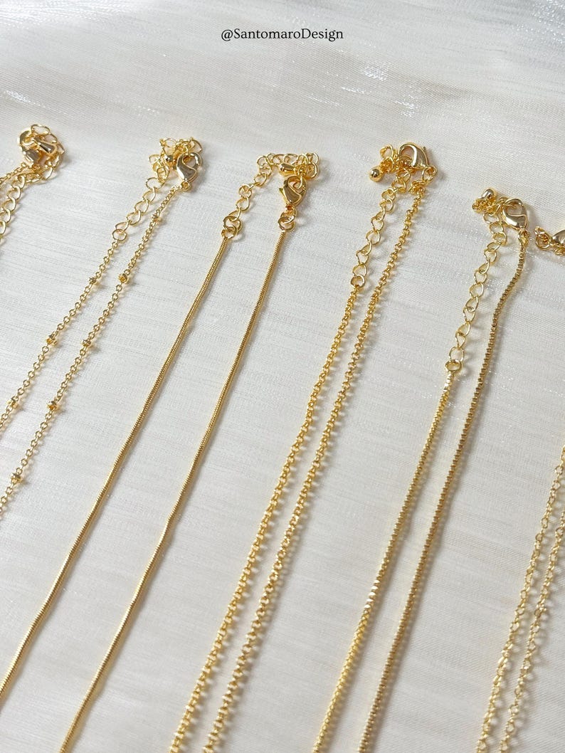 Op de afbeelding: Close-up van verschillende goudkleurige kettingen met diverse kettingstijlen. De kettingen hebben verschillende ontwerpen, waaronder kralen-, slang- en schakelkettingen. Elke ketting heeft een sluiting en een verstelbare ketting. De achtergrond is een witte, getextureerde stof.