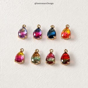 Puede incluir: Ocho colgantes en forma de lágrima con engastes dorados. Cada colgante presenta una gema facetada y colorida en tonos rosa, azul, amarillo y verde. Los colgantes están dispuestos sobre una superficie blanca.