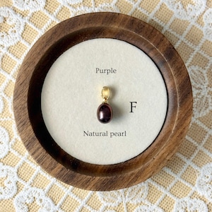 May include: A natural purple pearl pendant with a gold bail, displayed in a round wooden frame. The text "Purple" and "Natural pearl" are visible. The pendant is on a white background.