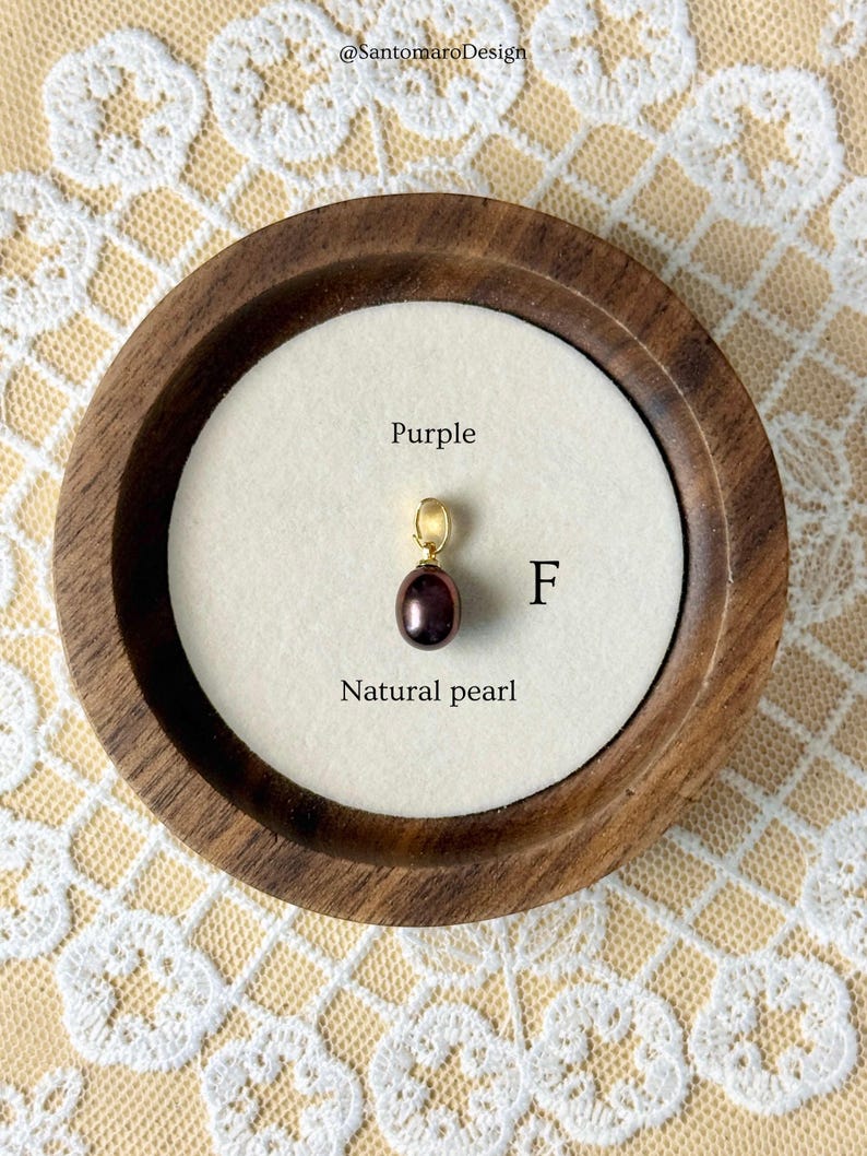 May include: A natural purple pearl pendant with a gold bail, displayed in a round wooden frame. The text "Purple" and "Natural pearl" are visible. The pendant is on a white background.