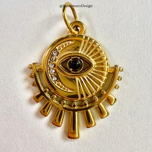 May include: Gold-coloured pendant with an eye design. The pendant features a black centre, a crescent moon, and a sunburst design. The eye is surrounded by a circle of small, clear crystals. The pendant has a loop at the top for a chain.