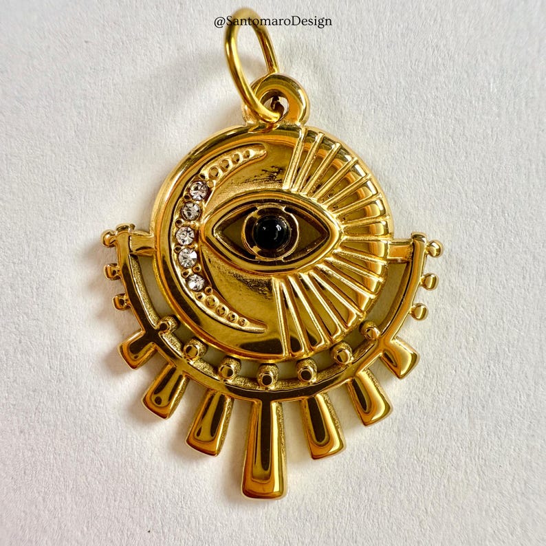 May include: Gold-coloured pendant with an eye design. The pendant features a black centre, a crescent moon, and a sunburst design. The eye is surrounded by a circle of small, clear crystals. The pendant has a loop at the top for a chain.
