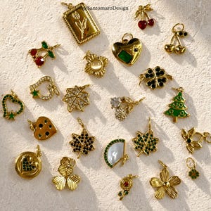 Può includere: Una collezione di ciondoli dorati con vari disegni, tra cui ciliegie, cuori, trifogli e fiori. Alcuni ciondoli presentano accenti verdi o neri, e altri hanno gemme chiare o bianche. I ciondoli sono disposti su una superficie strutturata bianco sporco.