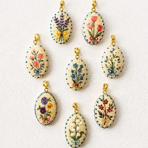 Può includere: Sette ciondoli ovali in ceramica con bouquet di fiori dipinti a mano su sfondo color crema. Ogni ciondolo presenta una contromaglia in metallo dorato e un bordo blu smerlato o puntinato. Il testo @SantomaroDesign è visibile in alto al centro.