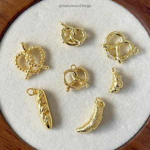 Op de afbeelding: Goudkleurige bedels in verschillende voedselvormen, waaronder pretzels, een croissant en een baguette. De bedels worden gepresenteerd op een wit oppervlak, met de tekst "@SantomaroDesign" zichtbaar in de linkerbovenhoek.