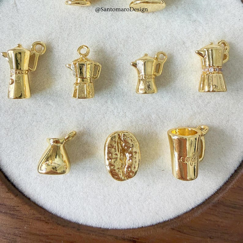 Może przedstawiać: Kolekcja złotych zawieszek o tematyce kawowej. Zawieszki obejmują miniaturowe ekspresy do kawy, ziarno kawy i kubek do kawy. Obraz zawiera tekst "@SantomaroDesign".