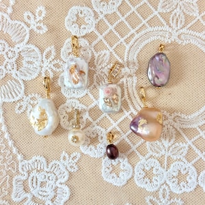 May include: A selection of gold pendants featuring pearls and gemstones. The designs include baroque pearls, a small pearl with a diamond accent, and a dark purple gemstone. The background is a cream-coloured lace.