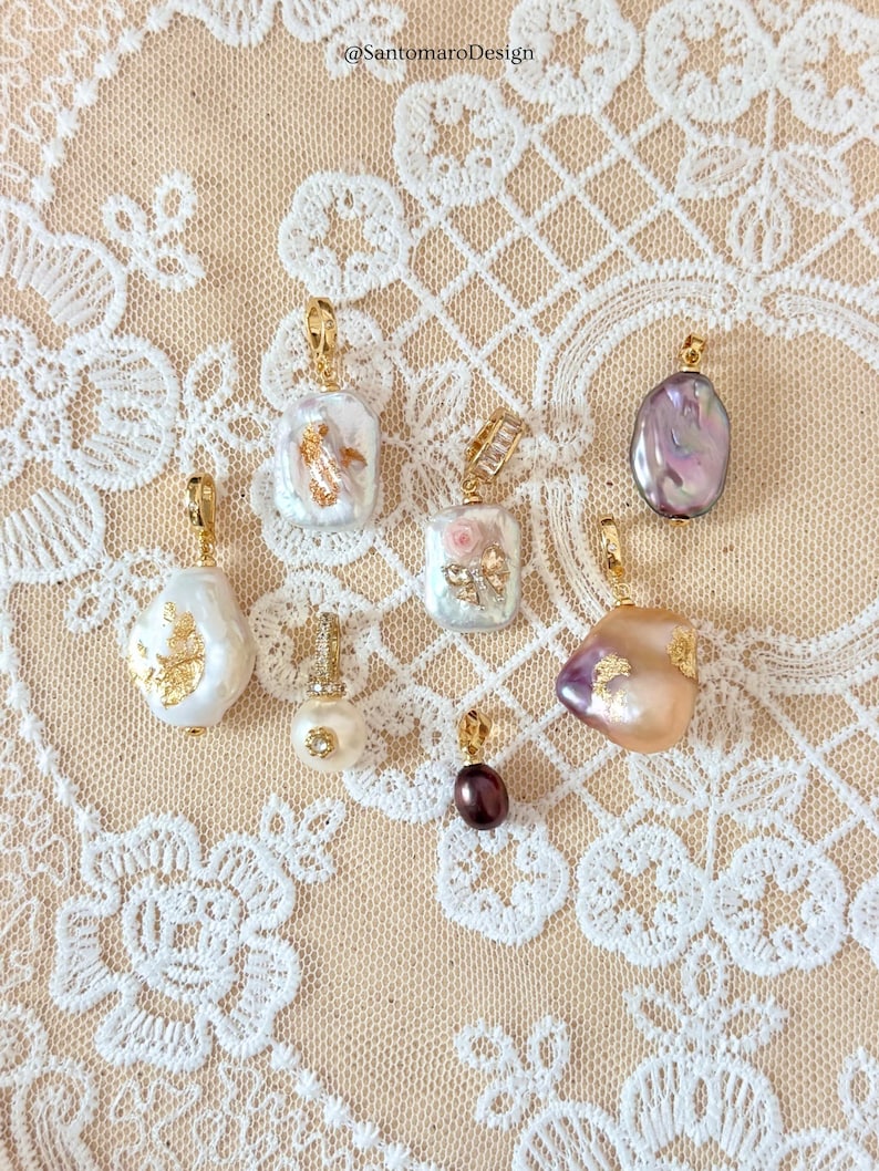May include: A selection of gold pendants featuring pearls and gemstones. The designs include baroque pearls, a small pearl with a diamond accent, and a dark purple gemstone. The background is a cream-coloured lace.