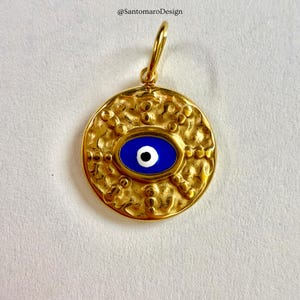May include: Gold-coloured pendant with a textured surface and a blue eye design. The pendant features a central oval with a blue background and a white and black eye detail. It has a loop at the top for hanging.