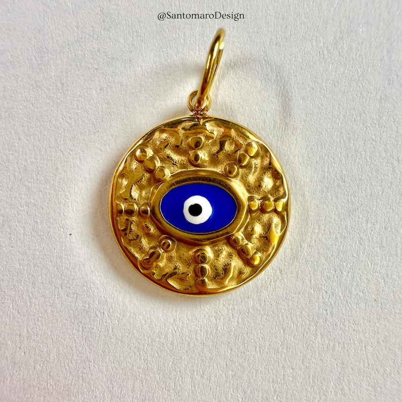 May include: Gold-coloured pendant with a textured surface and a blue eye design. The pendant features a central oval with a blue background and a white and black eye detail. It has a loop at the top for hanging.