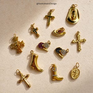 May include: A selection of gold-coloured charms, including crosses, cowboy boots, a cowboy hat, and heart-shaped pendants. Some charms are embellished with gemstones. The charms are displayed on a light-coloured surface.