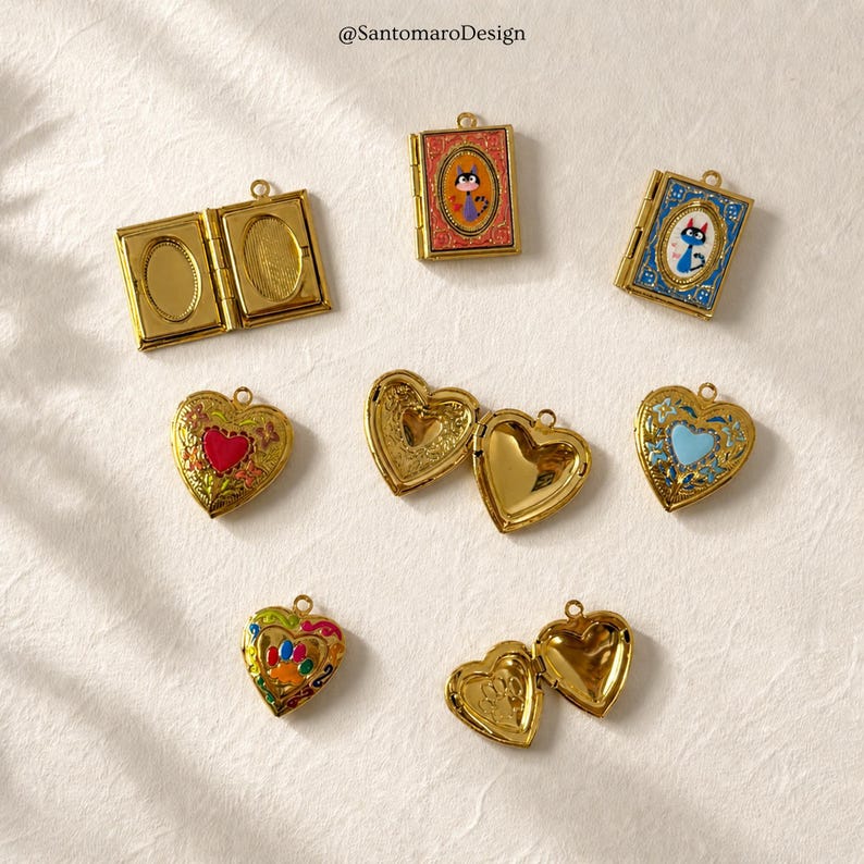 K&ouml;nnte beinhalten: Eine Sammlung goldfarbener Medaillons in verschiedenen Formen, darunter Herz- und Buchdesigns. Einige Medaillons sind ge&ouml;ffnet und zeigen das Innere. Mehrere weisen farbenfrohe Emaille-Details auf, wie ein rotes Herz, ein blaues Herz und eine Katzenillustration. Der Text "@SantamaroDesign" ist sichtbar.