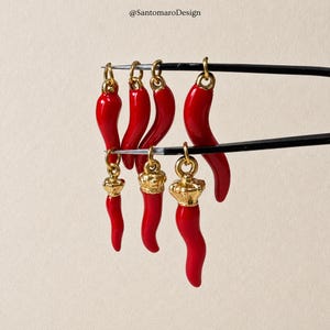 Könnte beinhalten: Eine Ansammlung von sechs roten Chilischoten-Anhängern, gehalten von einer schwarzen Pinzette. Jeder Anhänger hat einen glänzend roten, geschwungenen Körper mit einer goldfarbenen Metallöse. Drei Anhänger haben ein goldenes Kronendetail darüber.