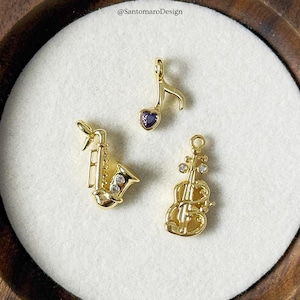 Pode incluir: Três pingentes dourados de instrumentos musicais: um saxofone, uma clave de sol com uma gema roxa em forma de coração e um violino com gemas transparentes. Os pingentes são exibidos em uma superfície branca.