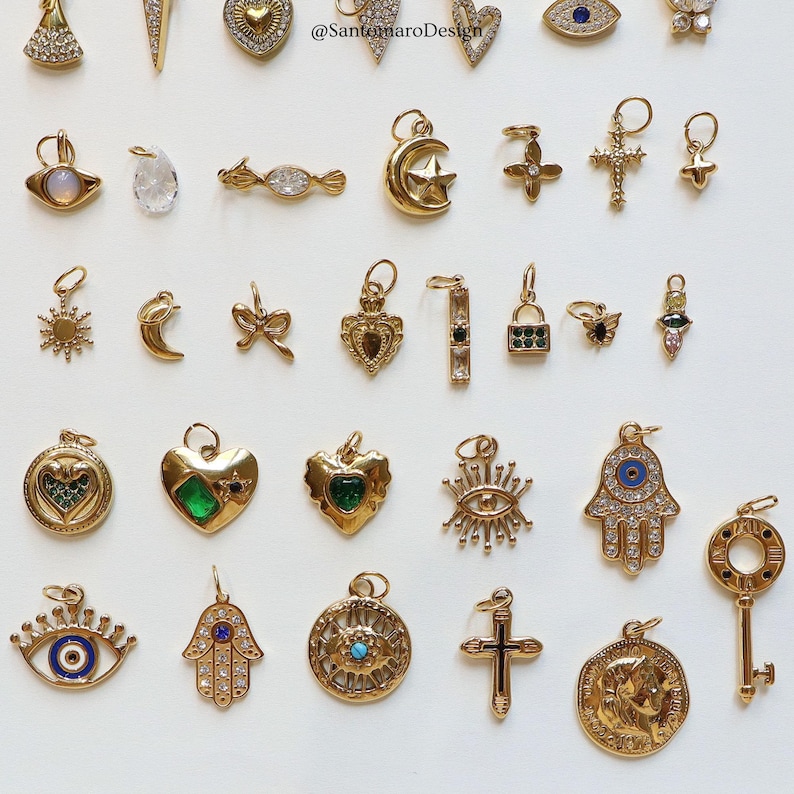May include: A collection of gold-colored charms with various designs, including hearts, crosses, eyes, and keys. Some charms feature gemstones and intricate detailing. The charms are displayed on a white surface.